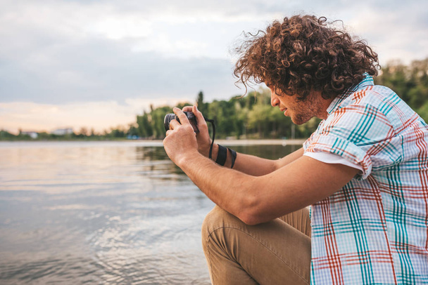 Side view of handsome male with curly hair checking photos of nature on his digital camera. Young handsome man wears casual shirt, with digital camera posing in the park. People, travel, lifestyle - Fotografie, Obrázek