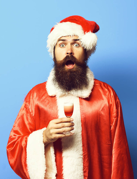 handsome bearded santa claus man with long beard on surprised face holding glass of alcoholic shot in red christmas or xmas sweater and new year hat on blue studio background - Photo, image