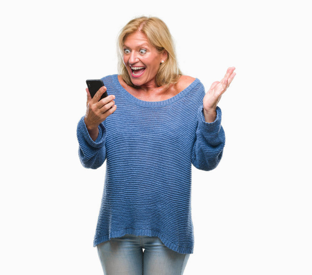 Blond žena středního věku odesílání zpráv pomocí smartphone přes samostatný pozadí velmi šťastný a nadšený, vítěz výraz slaví vítězství křičet s velkým úsměvem a pozvedl ruce - Fotografie, Obrázek