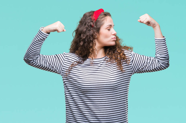 Beautiful brunette curly hair young girl wearing stripes sweater over isolated background showing arms muscles smiling proud. Fitness concept. - Fotoğraf, Görsel