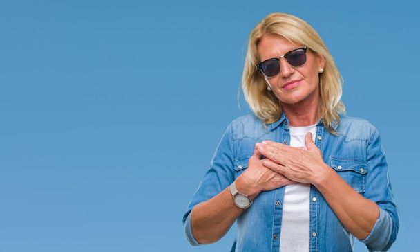 Middle age blonde woman wearing sunglasses over isolated background smiling with hands on chest with closed eyes and grateful gesture on face. Health concept. - Photo, Image