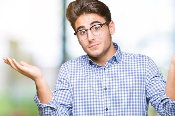Young handsome man wearing glasses over isolated background clueless and confused expression with arms and hands raised. Doubt concept. - Photo, Image