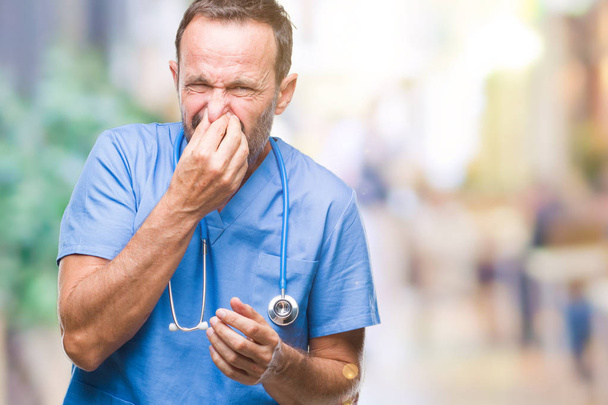 Middle age hoary senior doctor man wearing medical uniform over isolated background smelling something stinky and disgusting, intolerable smell, holding breath with fingers on nose. Bad smells concept. - Photo, Image