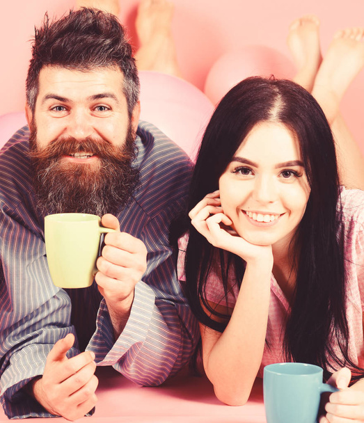 Man and woman on smiling faces lay, pink background. Couple in love drink coffee in bed. Man and woman in domestic clothes, pajamas. Couple relax in morning with coffee. Family tradition concept - Foto, Imagen