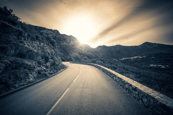 Musta ja valkoinen kuva kohti aurinkoa, kun se laskee mutkaiselle tielle Balagnen alueen vuoristossa Korsikalla
 - Valokuva, kuva