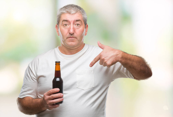 schöner älterer Mann trinkt Bierflasche über isoliertem Hintergrund mit überraschendem Gesicht, das mit dem Finger auf sich selbst zeigt - Foto, Bild