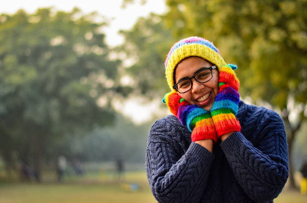 Mujer india joven con gorra de lana colorida, guantes, mitones y suéter azul, cubriéndose la cara con las manos y sonriendo en un parque en Delhi, India
 - Foto, imagen