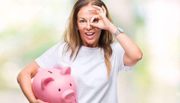 Латиноамериканка среднего возраста экономит деньги, используя копилку на изолированном фоне со счастливым лицом, улыбаясь, делая хорошо знак с рукой на глазу, глядя сквозь пальцы
 - Фото, изображение