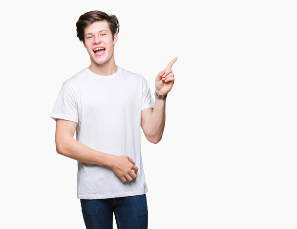 junger gutaussehender Mann in lässigem weißem T-Shirt vor isoliertem Hintergrund mit einem breiten Lächeln im Gesicht, der mit der Hand und dem Finger zur Seite in die Kamera zeigt. - Foto, Bild