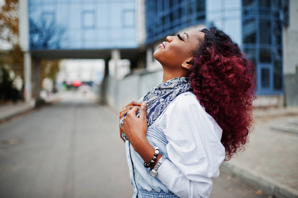 Attractive curly african american woman in jeans dress posed against modern multistory building. - Foto, Bild