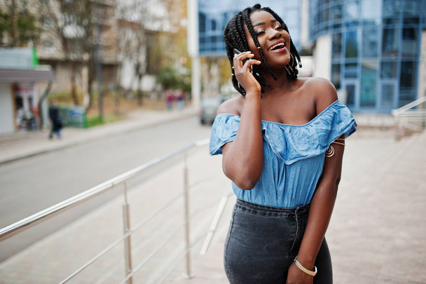 Attractive african american woman with dreads in jeans wear posed near railings against modern multistory building speaking at mobile phone. - Photo, Image
