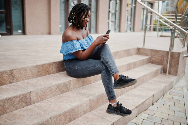 Attractive african american woman with dreads in jeans wear sitting on stairs with mobile phone. - Фото, изображение