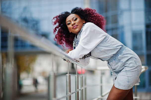 Attractive curly african american woman in jeans dress posed against modern multistory building. - Valokuva, kuva