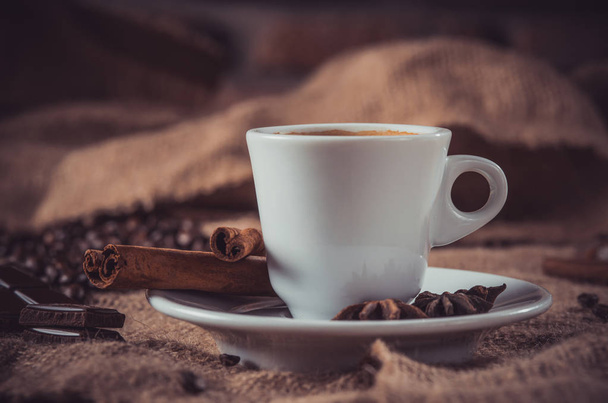camino oscuro de taza blanca de café con leche y especias aromáticas: palitos de canela, anís estrellado, rodajas secas de limón y granos de café en una servilleta de arpillera
 - Foto, Imagen