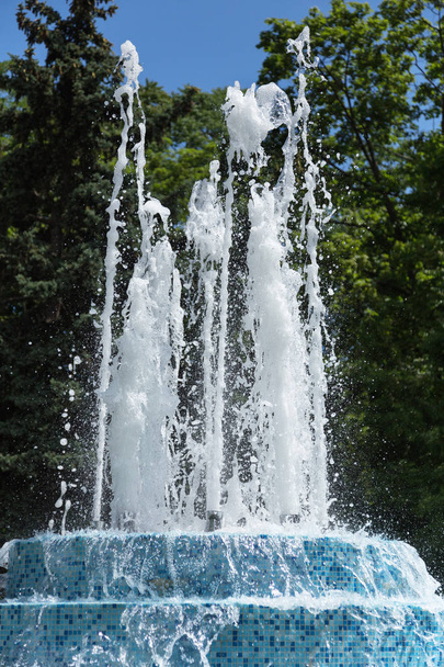 City fountain. Fountain in city park on a hot summer day. Stream of water, drops and bright splashes of water in beautiful city fountain. Bright streams of sparkling water in city fountain in day - Photo, Image