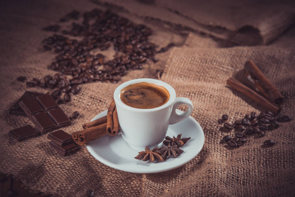 camino oscuro de taza blanca de café con leche y especias aromáticas: palitos de canela, anís estrellado, rodajas secas de limón y granos de café en una servilleta de arpillera
 - Foto, Imagen