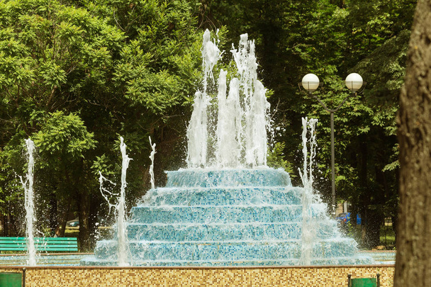 City fountain. Fountain in city park on hot summer day. Stream of water, drops and bright splashes of water in beautiful city fountain. Bright streams of sparkling water in city fountain in sun day - Photo, Image