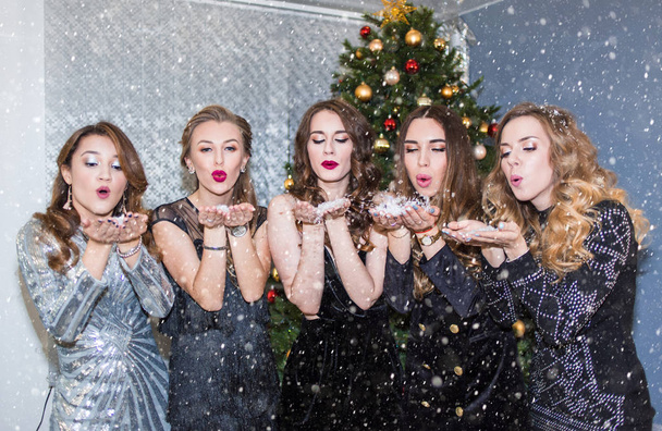 Beautiful young women having fun at a Christmas party, blowing away confetti and snow, sending kisses. New year, holiday, celebration, party - Foto, Imagem