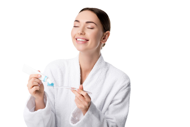 beautiful happy woman in white bathrobe putting toothpaste on toothbrush isolated on white - Valokuva, kuva