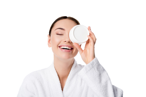 cheerful woman in bathrobe holding moisturizing cream in front of face isolated on white - Zdjęcie, obraz