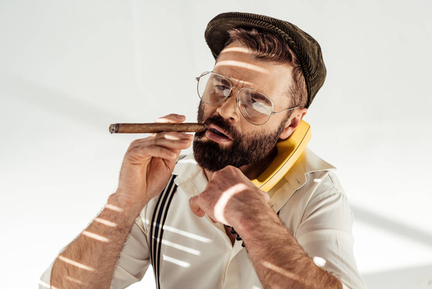 handsome man in glasses and cap talking on vintage phone and smoking cigar - Foto, Imagen