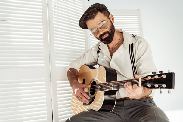 handsome bearded man in cap and glasses sitting and playing guitar near white room divider - Fotografie, Obrázek
