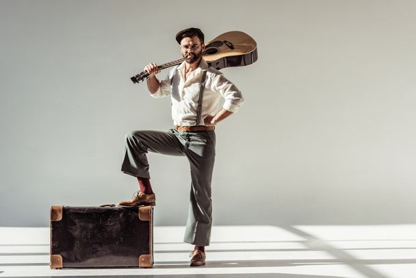 bearded man in cap with vintage suitcase holding acoustic guitar and looking at camera on grey background - Foto, afbeelding