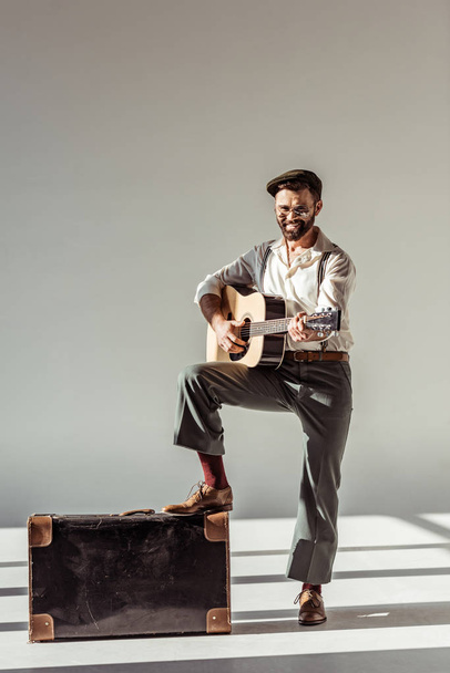 smiling bearded man in cap near vintage suitcase playing acoustic guitar and looking at camera on grey background - Fotoğraf, Görsel