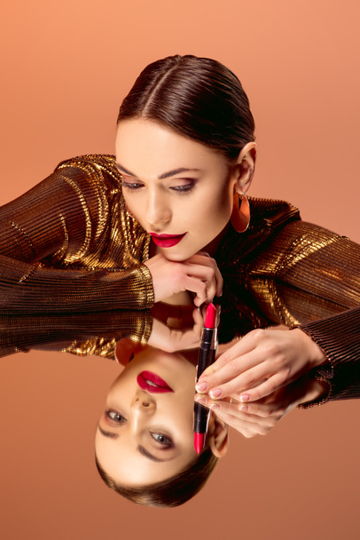 beautiful woman with glamorous makeup holding red lipstick and posing with mirror reflection isolated on orange - Фото, изображение