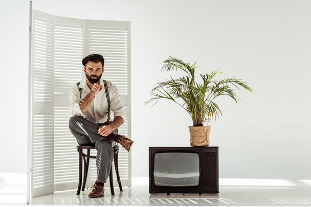 schöner bärtiger Mann sitzt auf Stuhl in der Nähe von Klappbildschirm und Fernseher im weißen Raum - Foto, Bild