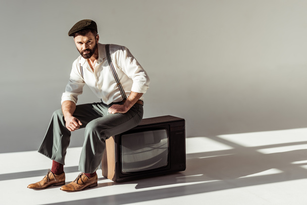 handsome stylish bearded man sitting on vintage tv and looking at camera - Foto, Imagen