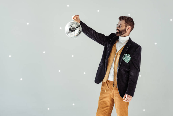 handsome bearded man looking at disco ball in hand on grey background - Φωτογραφία, εικόνα