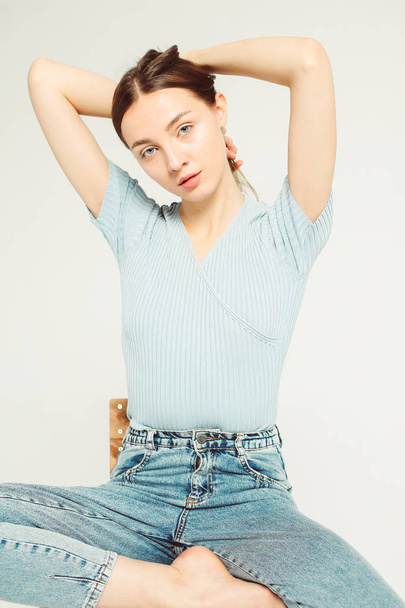 light portrait of pretty woman with dark hair, wearing blue t-shirt and jeans, is sitting on wooden chair, posing, touching her hair, hands over head, isolated in studio, grey background - 写真・画像