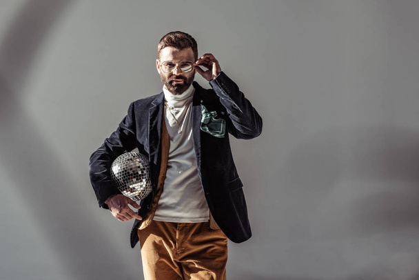 bearded man touching glasses, holding mosaic disco ball and looking at camera on grey background - Φωτογραφία, εικόνα