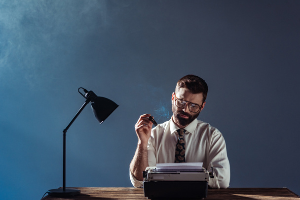 periodista barbudo sentado en la mesa con lámpara, mirando la máquina de escribir vintage y fumando sobre fondo gris
 - Foto, Imagen