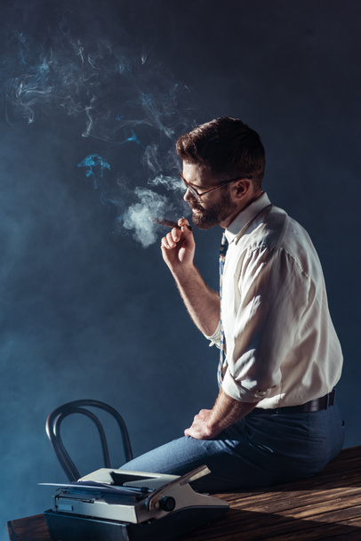 handsome bearded man in glasses sitting on table while smoking cigar - Photo, Image