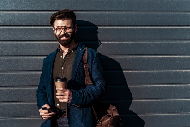 handsome bearded man in glasses and cap holding paper cup and smiling at camera - Фото, зображення