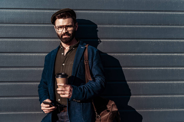 handsome bearded man in glasses and cap holding paper cup and smartphone - Fotó, kép
