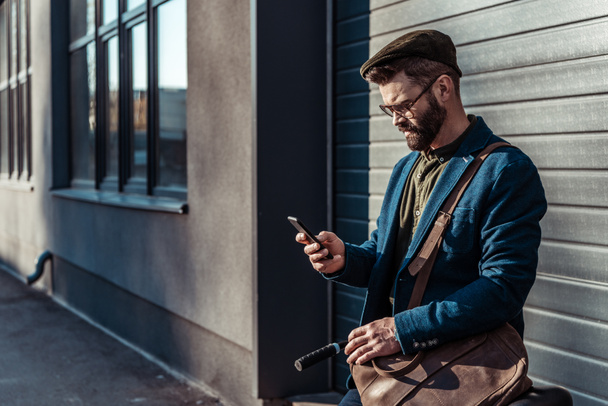 handsome bearded man in glasses and cap holding bicycle and looking at smartphone - Photo, Image