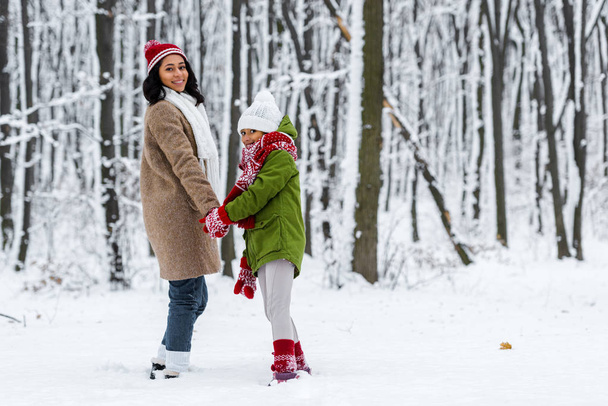 アフリカ系アメリカ人の母と娘、笑みを浮かべて、手を繋いでいると冬の公園でカメラを見て - 写真・画像