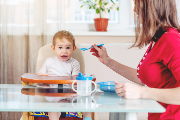 Мама кормит ребенка, протягивая руку ложкой еды на кухне. Здоровое детское питание. Эмоции ребенка во время еды
 - Фото, изображение