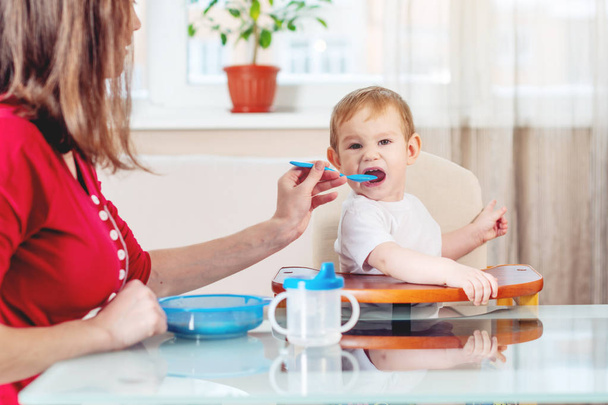 Mom feeding the baby holding out her hand with a spoon of food in the kitchen. Healthy baby food. The emotions of a child while eating - Photo, image