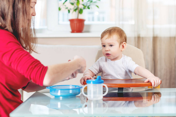 Мать кормит ребенка, протягивая руку ложкой каши на кухне. Эмоции ребенка во время еды здоровой пищи
. - Фото, изображение