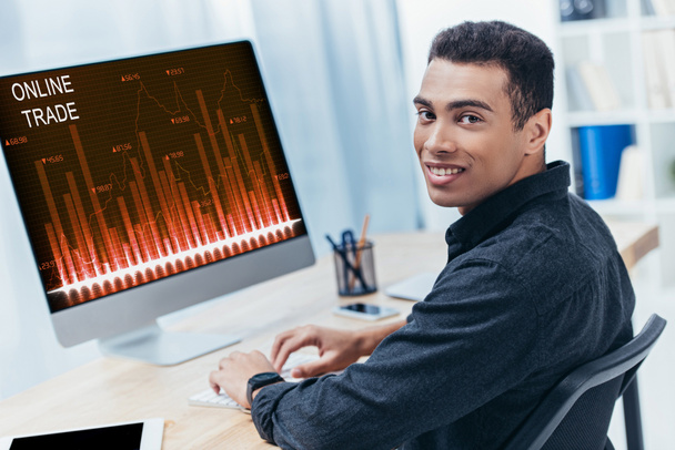mladí Smíšené rasy podnikatel pomocí stolního počítače s on-line obchodní grafy a usmívá se na kameru - Fotografie, Obrázek