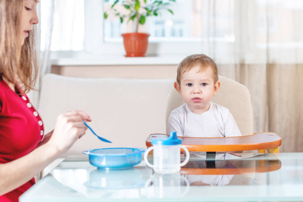 Mère nourrissant le bébé en lui tenant la main avec une cuillère de porridge dans la cuisine. Émotions d'un enfant en mangeant des aliments sains
. - Photo, image