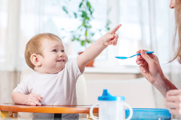 Mother feeding the baby holding out her hand with a spoon of porridge in the kitchen. Emotions of a child while eating healthy food. - Photo, Image