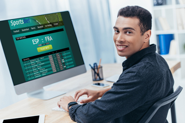 young mixed race businessman using desktop computer with sports bet website and smiling at camera   - Фото, зображення