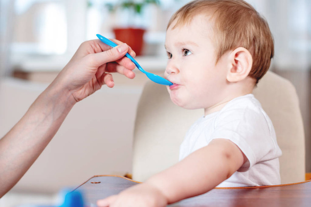Mother feeding the baby holding out her hand with a spoon of porridge in the kitchen. Emotions of a child while eating healthy food. - 写真・画像
