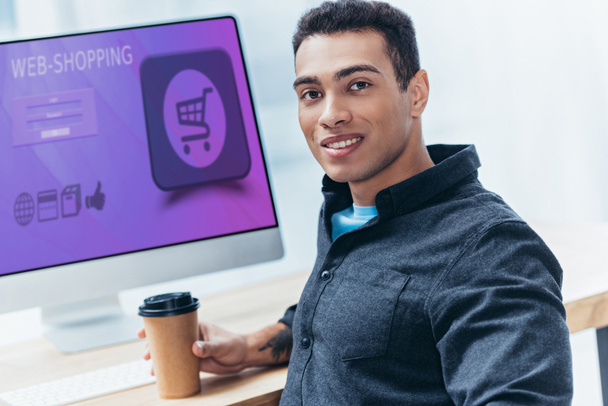 young businessman with coffee to go working with desktop computer with shopping website and smiling at camera - Photo, Image