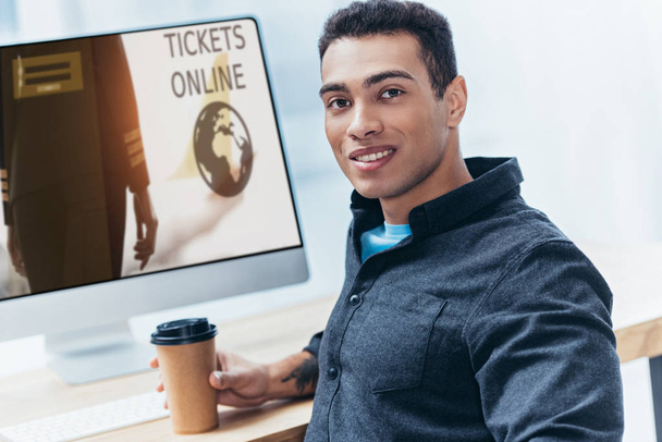 nuori liikemies, jolla on kahvia mennä työskentelemään pöytätietokoneen kanssa, jolla on liput online-sivustoon ja hymyilee kameralle
 - Valokuva, kuva